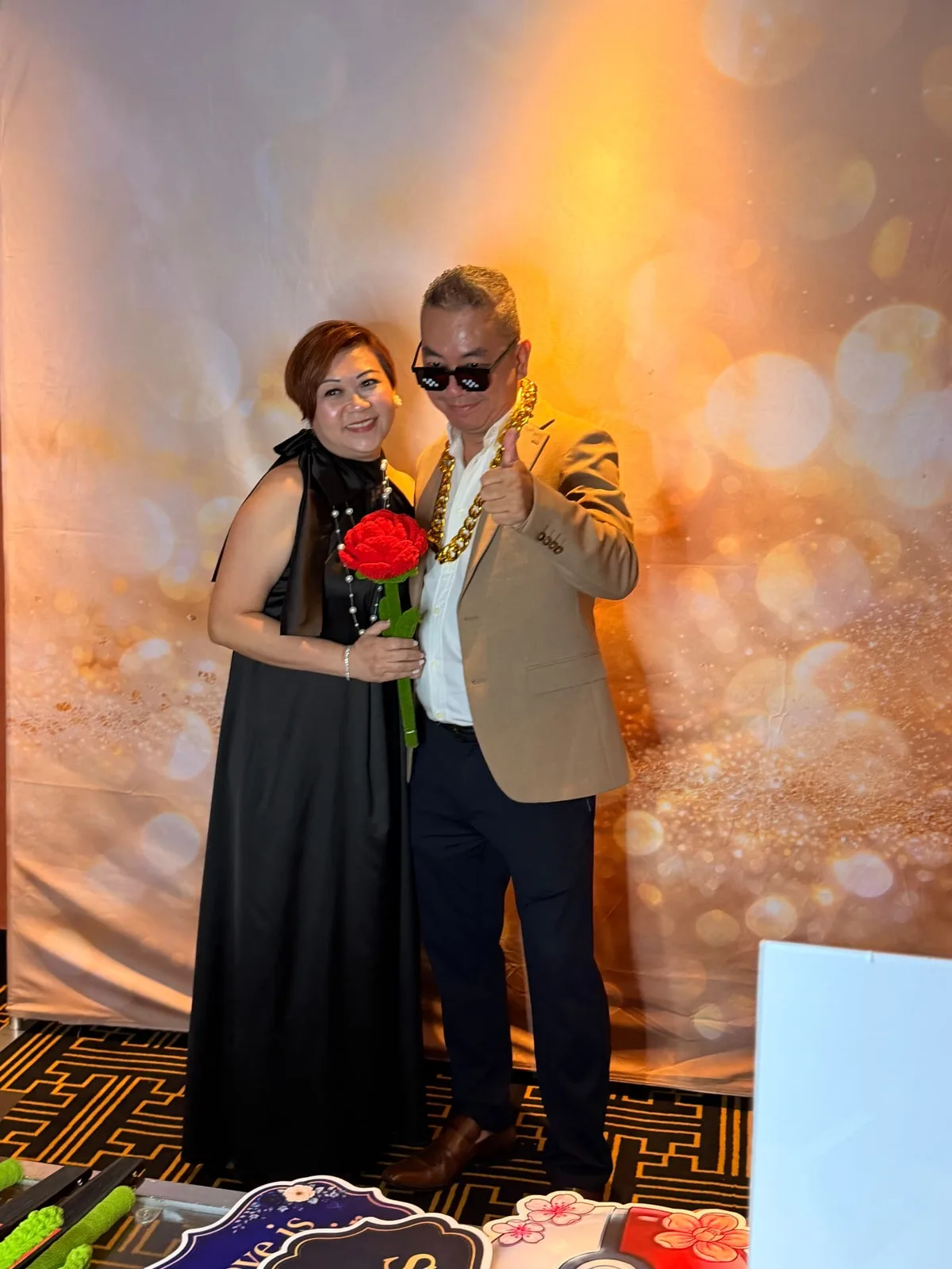 Wedding guests posing with a rose prop and party sunglasses at a gold photobooth backdrop in Singapore